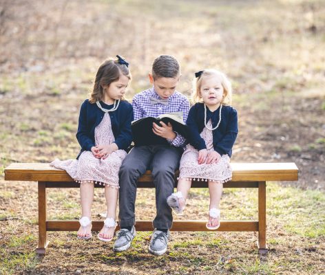 A little boy reading a book for two girls sitting on a bench in a park