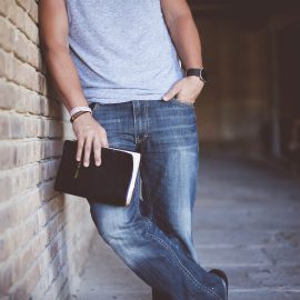 A closeup shot of a male leaning against a wall while holding the bible with a blurred background